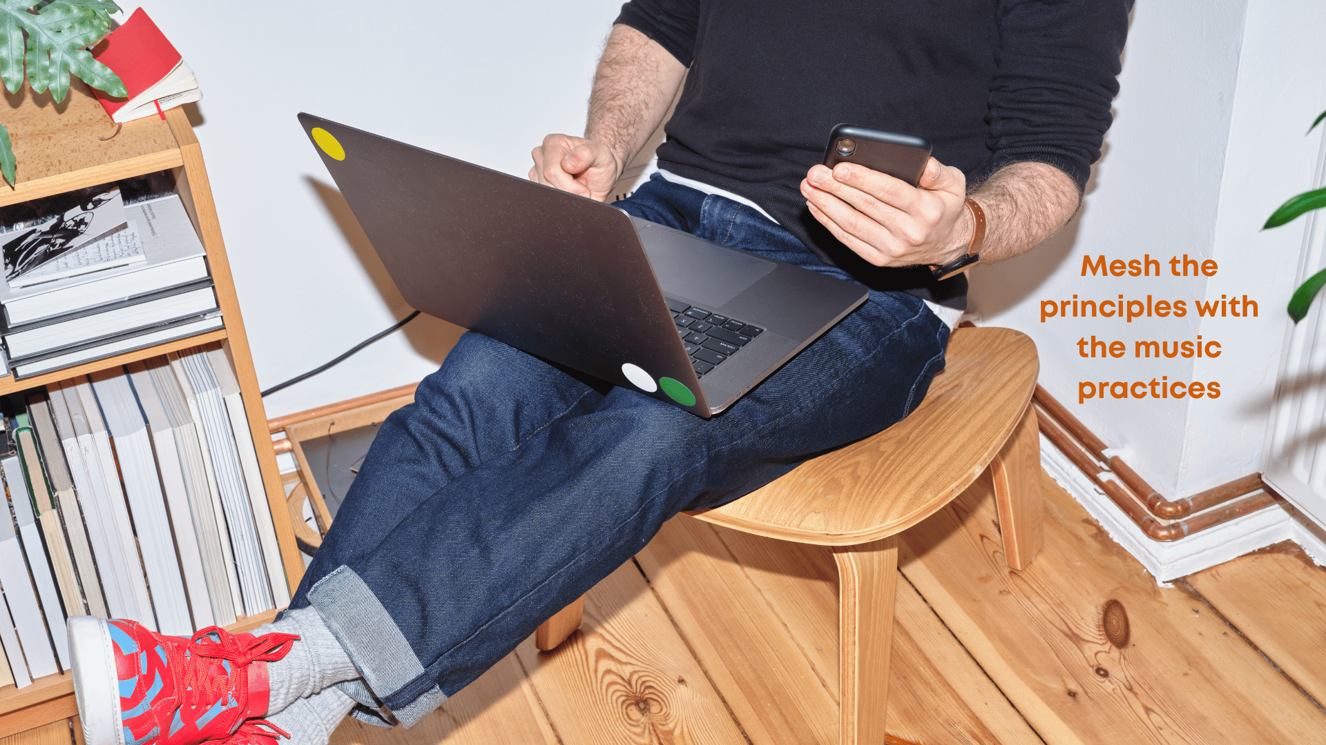 5. principles underpin practices A man wearing jeans and sneakers with red shoelaces relaxes on a low stool by a bookshelf, he holds a phone and a laptop.